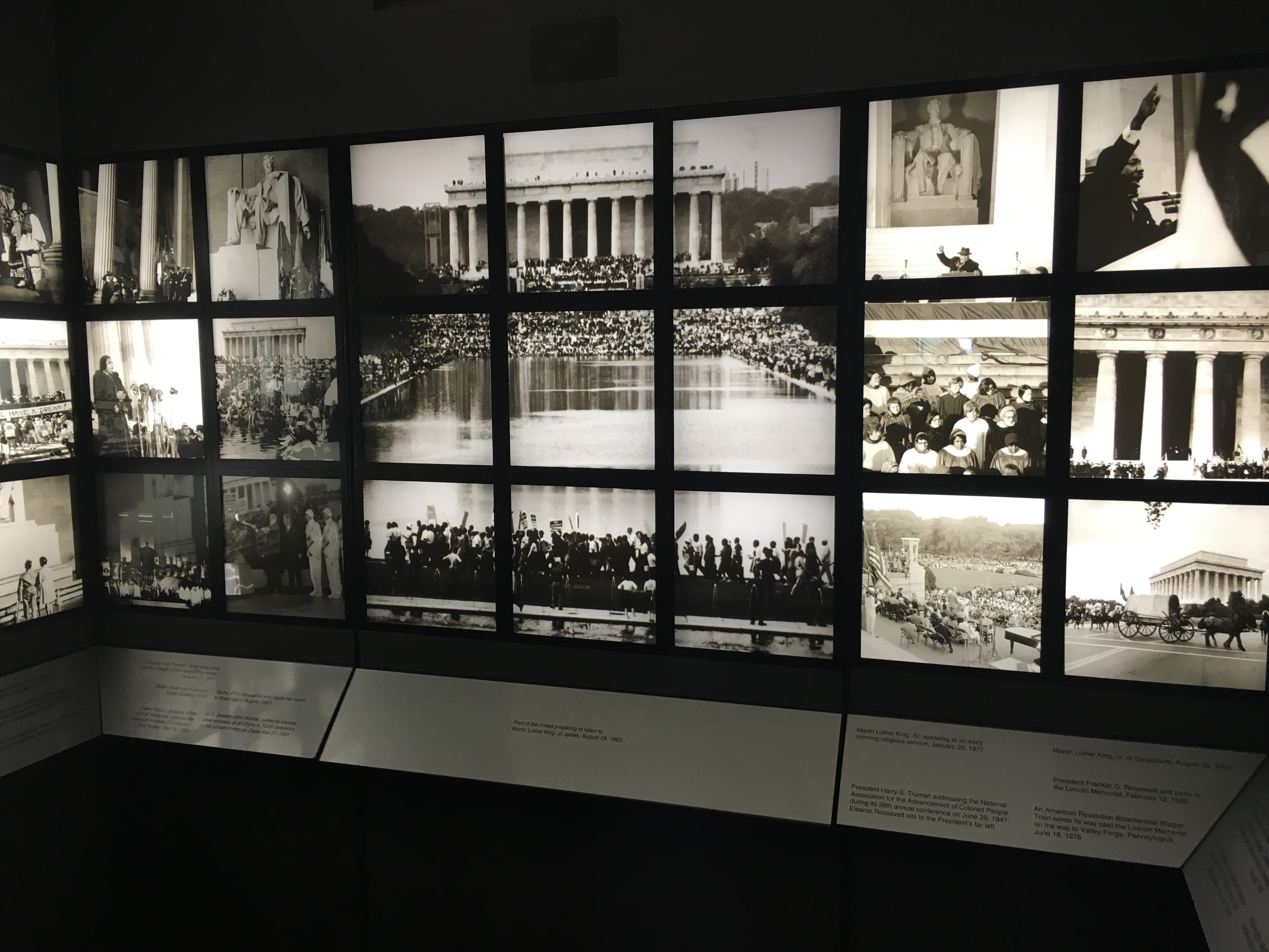 Museum exhibit wall displaying a grid of black-and-white historical photographs centered on the Lincoln Memorial and Reflecting Pool. The images show large public gatherings, speeches, ceremonial events, and crowds assembled around the memorial. Interpretive text panels run along the bottom of the display in a dimly lit gallery.