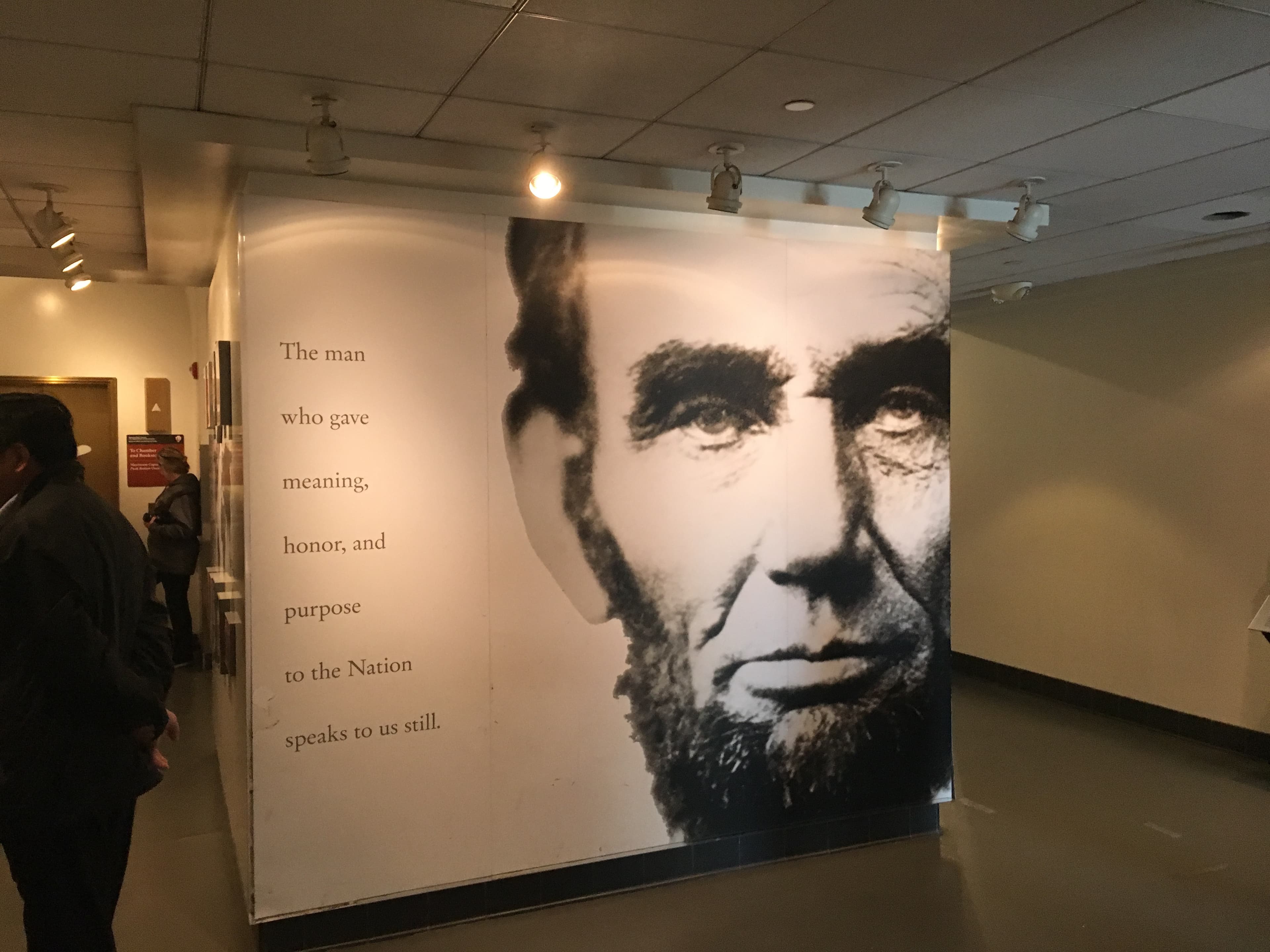 Interior museum display featuring a large black-and-white portrait of Abraham Lincoln printed across a corner wall. Beside the portrait, a quotation reads: "The man who gave meaning, honor, and purpose to the Nation speaks to us still." Visitors stand nearby in a softly lit exhibit space