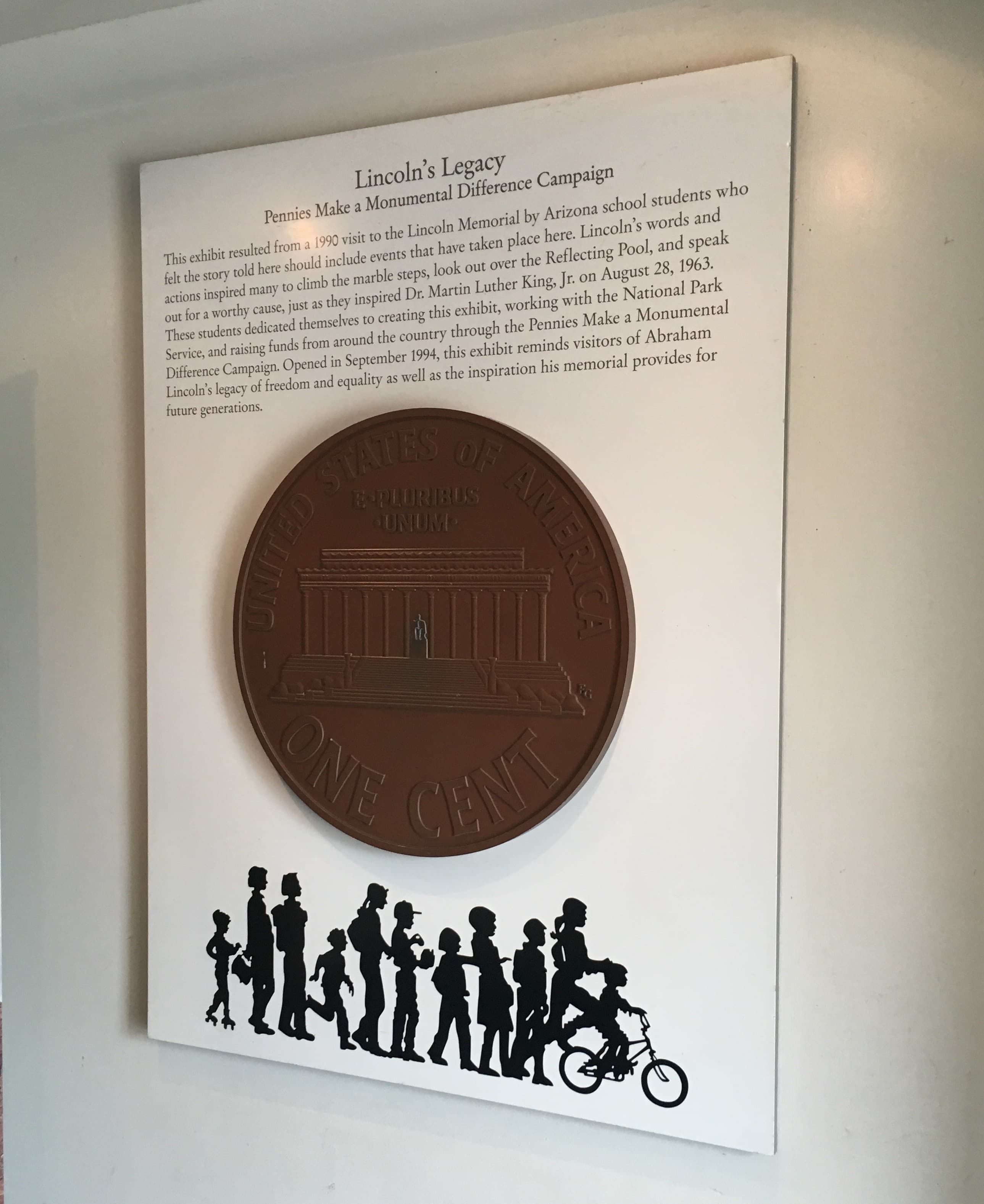 Museum exhibit panel titled “Lincoln’s Legacy: Pennies Make a Monumental Difference Campaign.” The display features an oversized replica of the reverse side of a U.S. penny showing the Lincoln Memorial, mounted beneath explanatory text about a student-led fundraising campaign. Along the bottom, black silhouettes of children and adults walking, standing, and riding a bicycle stretch across the panel.