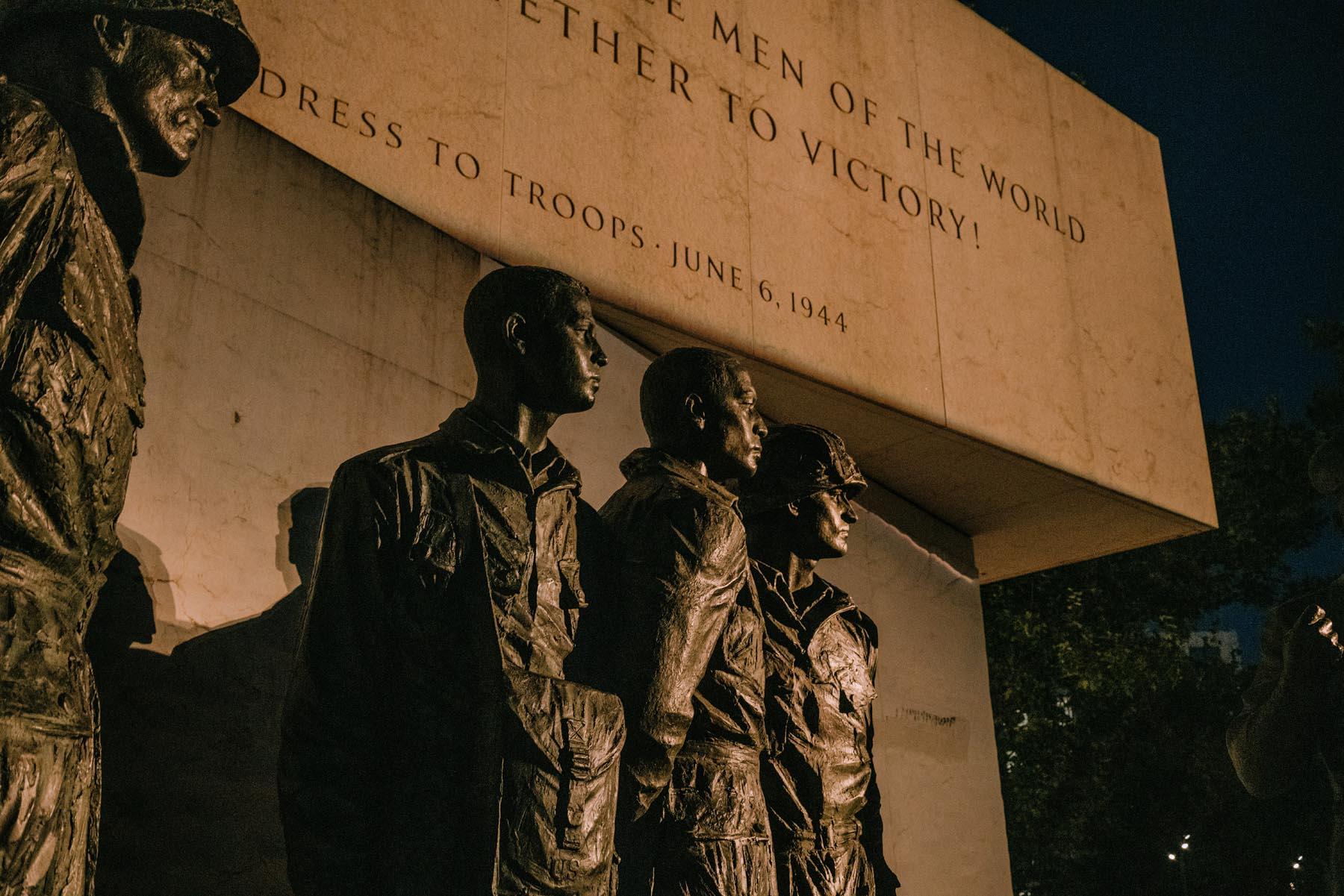 Close-up of bronze statues of soldiers, lit dramatically at night against the backdrop of Eisenhower's D-Day address engraved in stone.