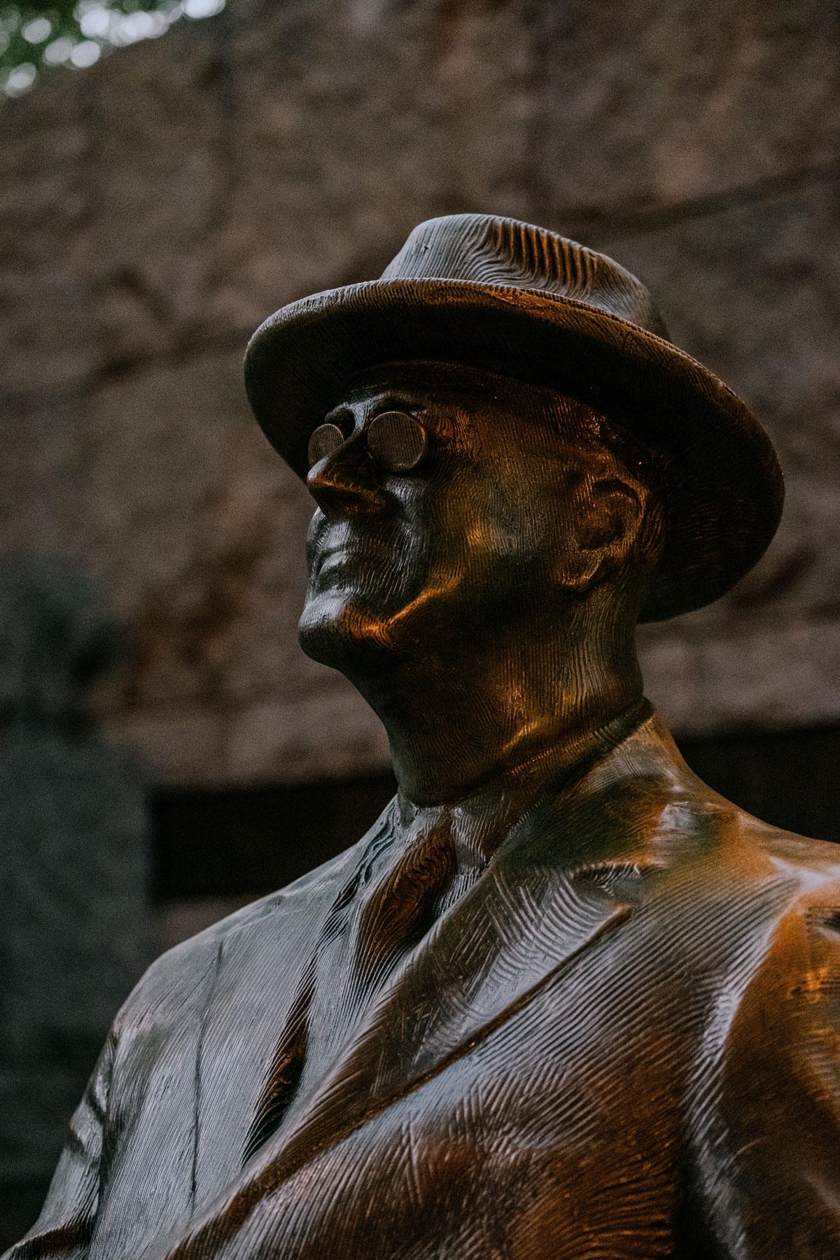 Close-up of Franklin D. Roosevelt’s statue wearing glasses and a hat, captured at dusk with textured details visible.