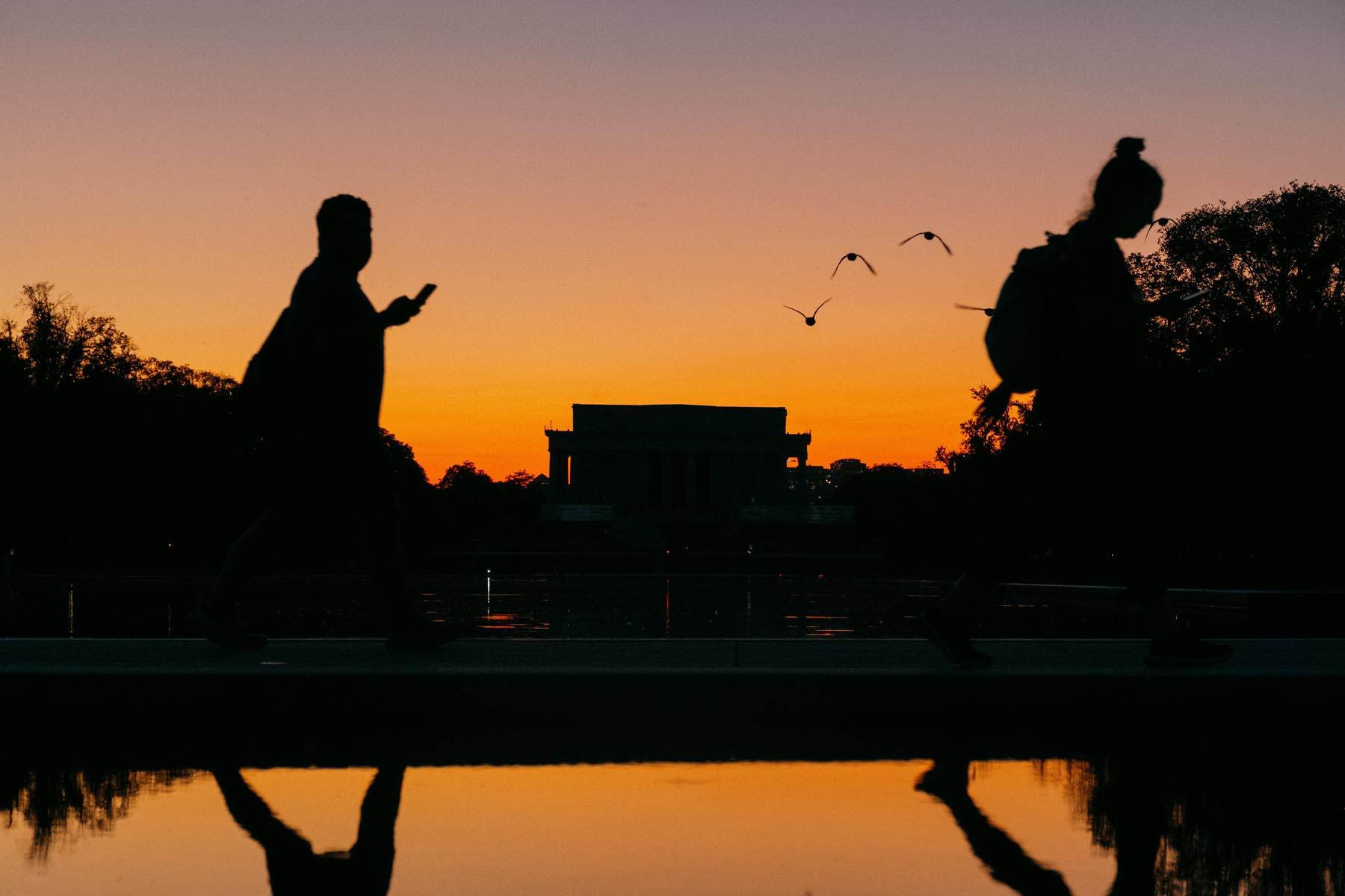 Silhouettes of two people walking near the Reflecting Pool at sunset with the Lincoln Memorial in the background, birds flying above, and the sky transitioning to shades of orange and purple.