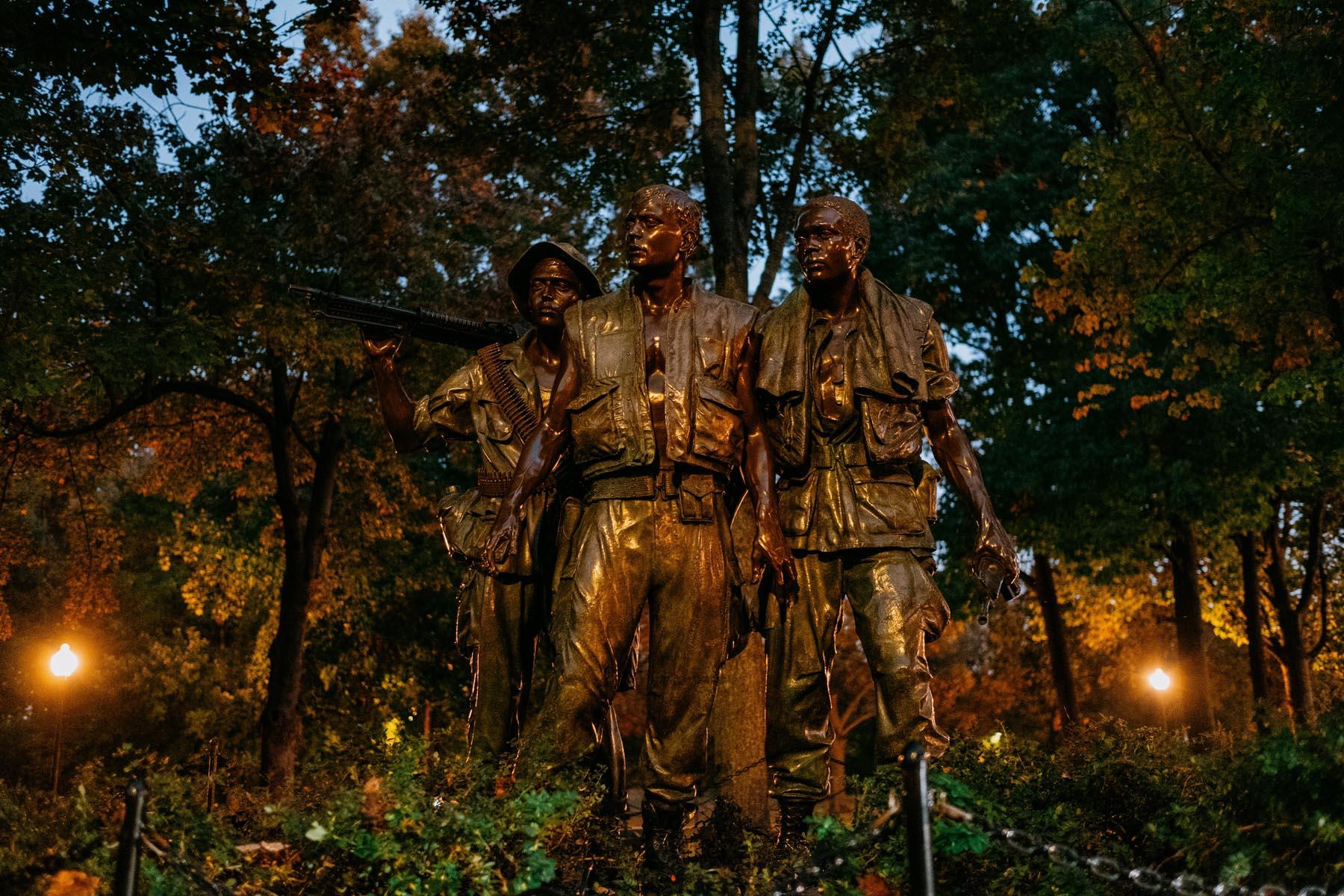 Wide-angle view of the Three Soldiers statue at night, emphasizing the trio’s formation and solemn expressions with soft lights in the background.