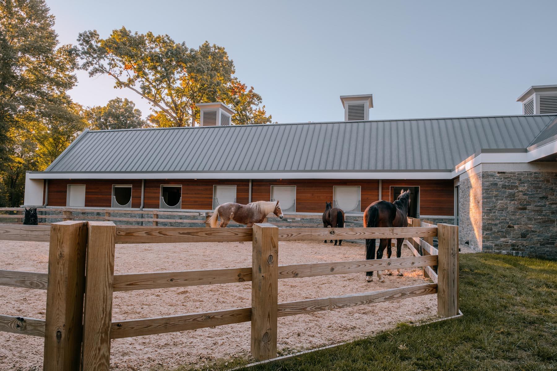 A wide view of the U.S. Park Police Horse Stables and Education Center, showcasing the wooden paddock, stable building, and horses grazing in the evening light.