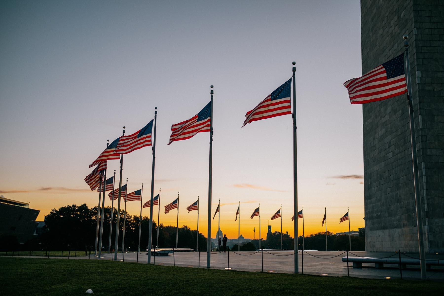 A line of American flags surrounding the Washington Monument at sunset, with a colorful sky and the U.S. Capitol faintly visible in the distance.