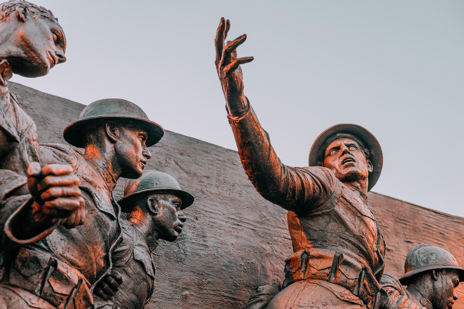 A group of bronze soldiers at the WWI memorial, with one leading with an outstretched arm, surrounded by comrades in helmets, illustrating camaraderie and leadership under dramatic lighting.